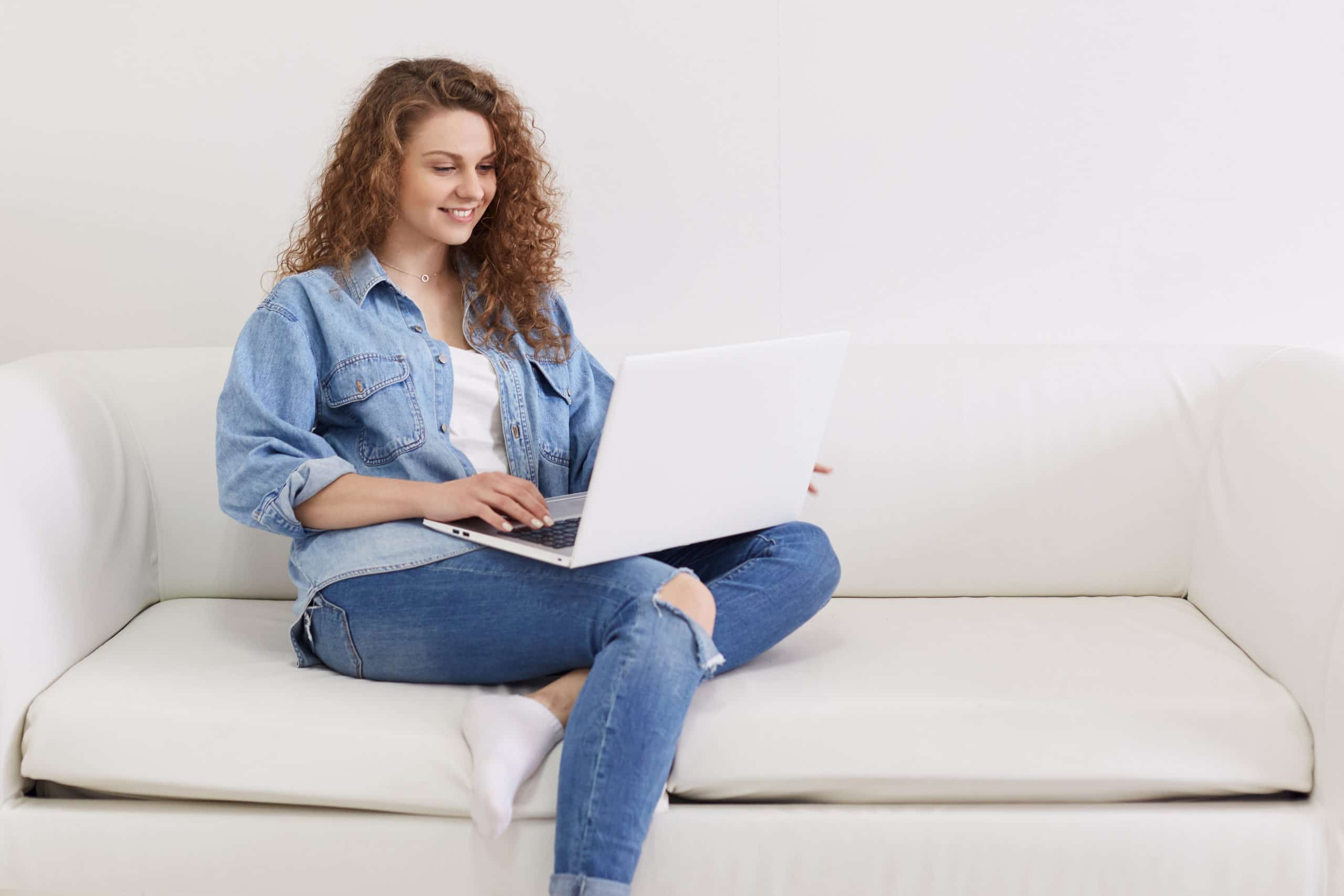 Horizontal shot of nice looking lovely attractive woman, focused curly haired girl sitting on white sofa while working remotely part time, using her modern portable computer and wireless Internet.